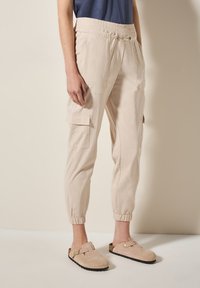 Pantalons cargo beiges avec poches latérales, taille élastique et chevilles resserrées. Associés à des sabots en suède beige avec une accent de boucle.