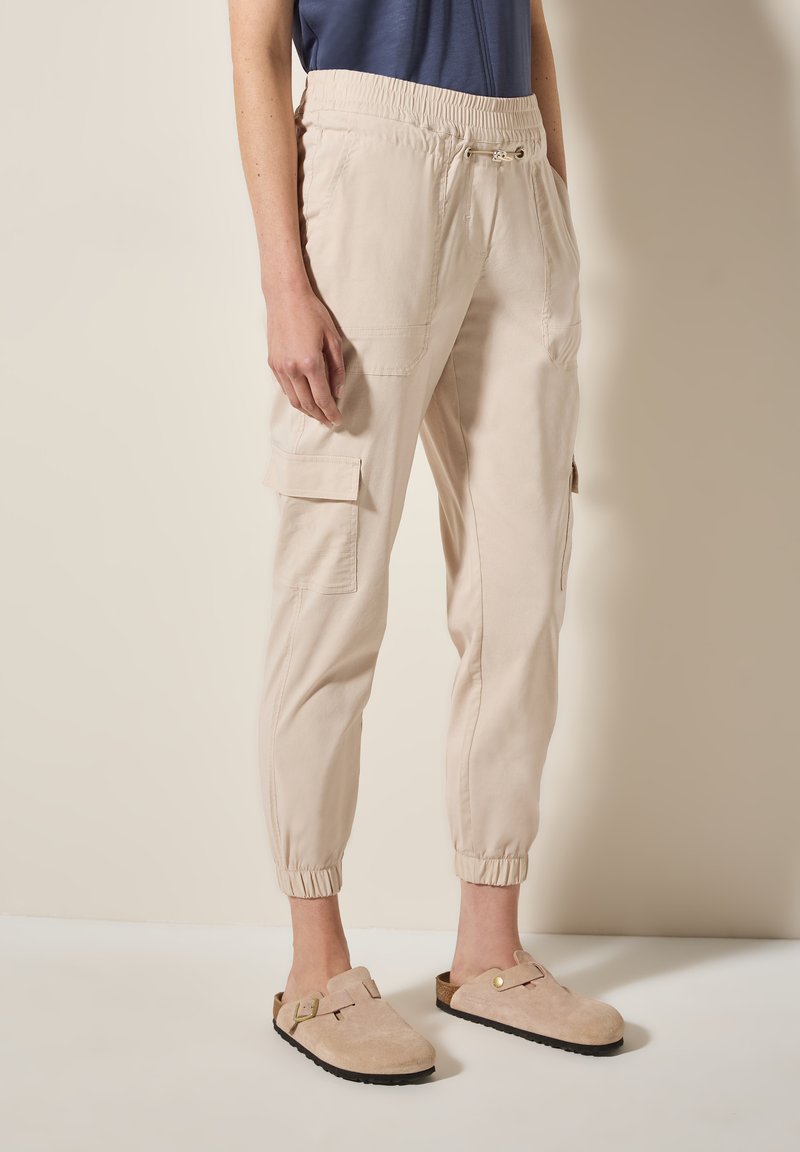 Pantalons cargo beiges avec poches latérales, taille élastique et chevilles resserrées. Associés à des sabots en suède beige avec une accent de boucle.