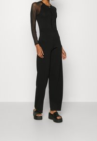 Bodysuit noir avec de longues manches transparentes et une fermeture éclair sur le devant, associé à un pantalon noir taille haute ample et des sandales à plateforme chunky.