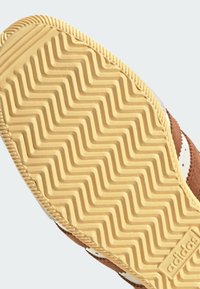 Brown suede sneaker sole with a zigzag rubber pattern, featuring white accents and textured edges. Logo visible on the side.