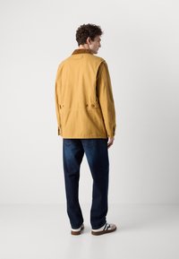Giacca marrone in tessuto senape, con dettagli a bottoni in vita, abbinata a jeans blu scuro e scarpe casual chiare.