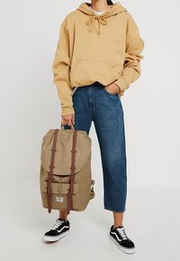 Khaki plecak z brązowymi paskami i składanym wierzchem, w połączeniu z beżową bluzą i niebieskimi dżinsami, noszony z czarnymi trampkami.