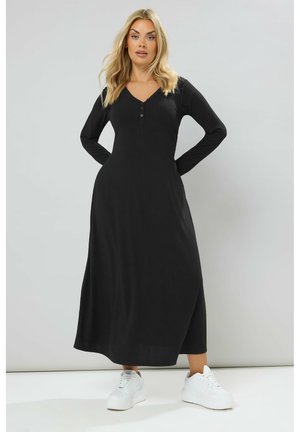 Schwarzes, geripptes Maxikleid mit langen Ärmeln und V-Ausschnitt, das Knopfakzente am Ausschnitt und eine fließende Silhouette aufweist. Trägt weiße Sneakers.