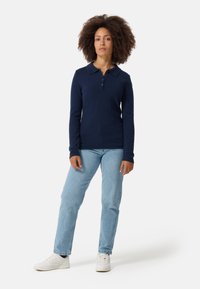 Granatowa koszula polo z długim rękawem, z kołnierzykiem i listwą na guziki, w połączeniu z jasnoniebieskimi dżinsami i białymi trampkami na jednolitym tle.