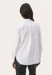 Weißes Langarmhemd mit Kentkragen, abgerundetem Saum und glatter Textur, kombiniert mit schwarzen Hosen. Minimalistisches Design ohne Muster.