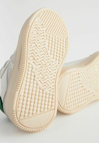 Semelles de baskets blanches avec des motifs en caoutchouc surélevés, comprenant des grips texturés et une base de couleur beige, présentant des motifs en zigzag et en points.