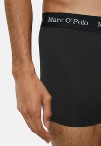 Musta puuvillainen bokserihousut pehmeällä, joustavalla vyötärönauhalla, jossa on valkoinen "Marc O'Polo" -logo. Käsi lepää sivulla.