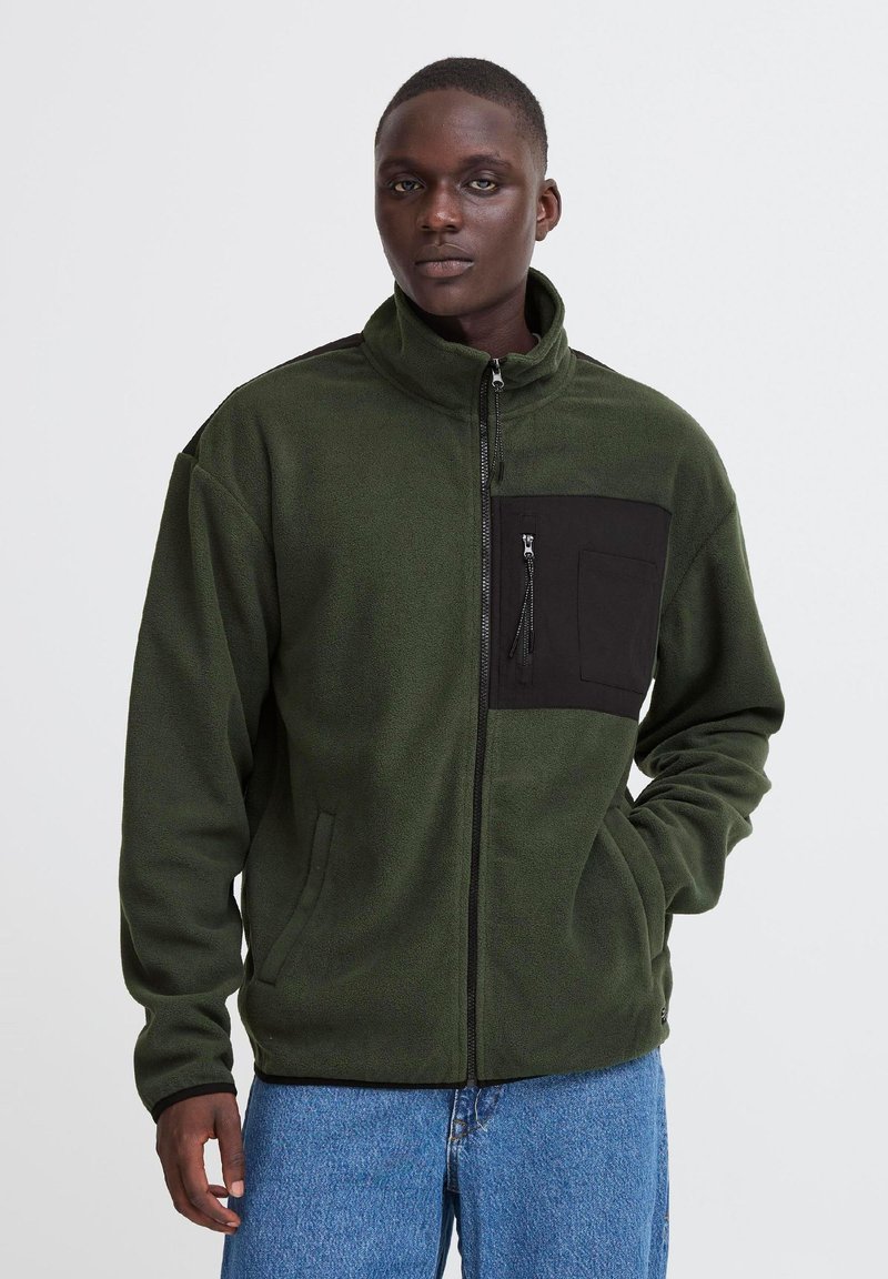 Personne portant une veste polaire zippée vert foncé avec une poche poitrine noire et un jean bleu, debout devant un fond uni clair.