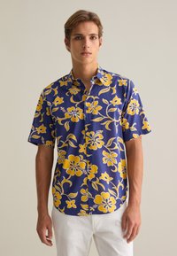 Camisa de manga corta con botones en color azul marino con grandes patrones florales amarillos. Hecha de una tela ligera, presenta un corte relajado y cuello.