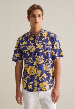 Camisa de manga corta con botones en color azul marino con grandes patrones florales amarillos. Hecha de una tela ligera, presenta un corte relajado y cuello.