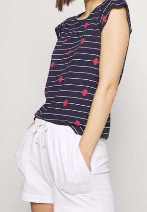 Kobieta ubrana w granatowy top z krótkim rękawem, z białymi paskami i czerwonymi nadrukami truskawek, w połączeniu z białymi szortami ze ściągaczem, ręce w kieszeniach.