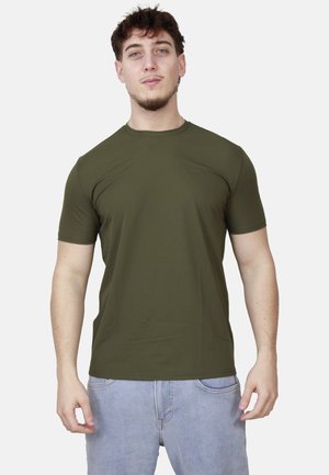 Giovane uomo in piedi di fronte, indossa una maglietta verde oliva a maniche corte e jeans azzurri chiari su uno sfondo uniforme.