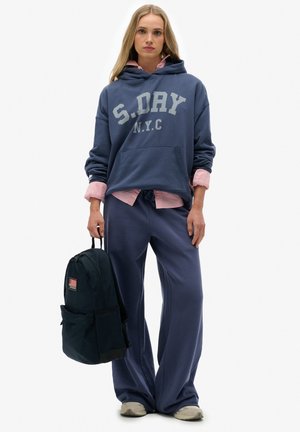 Jonge vrouw die een blauwe S.DRY NYC-hoodie draagt, een roze shirt, losse blauwe broek, grijze sneakers, en een zwarte rugzak naast zich houdt.