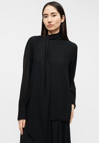 Μαύρη μπλούζα με μακριά μανίκια, ψηλό λαιμό και λεπτομέρεια που μοιάζει με φουλάρι. Κατασκευασμένη από ελαφρύ ύφασμα, διαθέτει μανσέτες με κουμπιά και άνετη γραμμή.