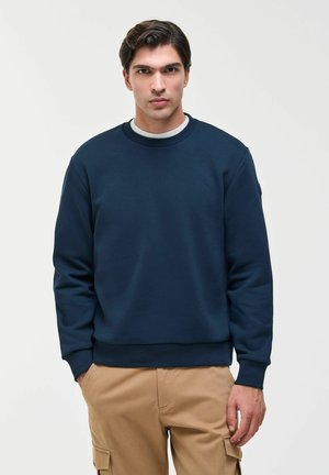 Giovane uomo con capelli scuri che indossa una felpa blu navy e pantaloni cargo beige, in piedi contro uno sfondo chiaro e semplice.