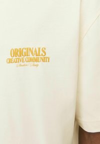Krēmpadējā T-krekls ar dzeltenu izšuvumu "ORIGINALS CREATIVE COMMUNITY", īsām piedurknēm un brīvu piegriezumu.