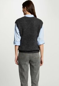 Gilet grigio in lana con scollatura rotonda, maniche corte e orlo inferiore aderente, abbinato a una camicia azzurro chiaro con maniche arrotolate e pantaloni in denim grigio.