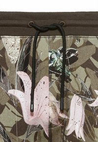 Tissu à motif camouflage en nuances de vert et de brun, avec des motifs floraux roses. Comprend une ceinture élastique marron avec un cordon noir.