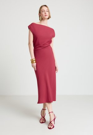 Donna che indossa un abito midi rosso senza maniche e con spalle scoperte, abbinato a tacchi rossi con cinturini e gioielli dorati, in piedi davanti a uno sfondo semplice.