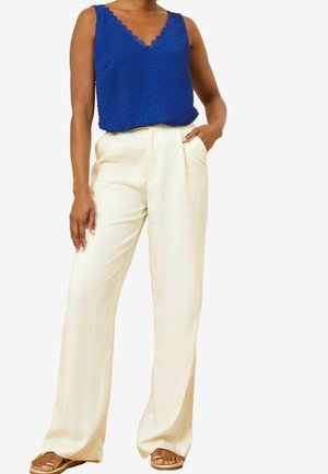 Femme portant un haut bleu sans manches texturé et un pantalon ample crème taille haute avec des sandales, main dans la poche, sur fond blanc.