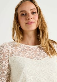 Blusa de encaje blanco con un patrón geométrico, con un escote redondo y mangas largas transparentes. Textura suave y diseño ligero y aireado.