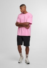 Mees seisab käed risti, seljas on tal lahtine roosa T-särk, mustad lühikesed püksid, beežid sokid ja hõbedased spordijalatsid, ühtlasel taustal.