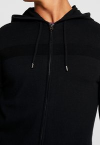 Black zip-up hoodie with a textured knit pattern, featuring a hood, drawstrings, and silver zipper accents.