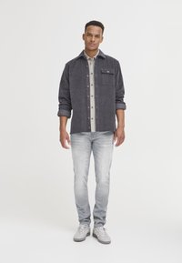 Grå kordflodskjorta med knäppning och en bröstficka, lagd över en randig skjorta, tillsammans med ljus tvättade jeans och grå sneakers.