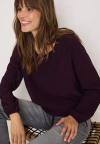 Blouse bourgogne avec des rayures verticales, col rond, et manches longues avec des poignets élastiques, associée à un jean gris clair.