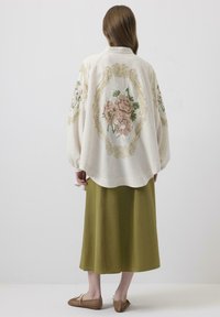 Bluză crem brodată, cu modele florale și accente aurii, asortată cu o fustă midi verde și sandale din piele împletită maro.
