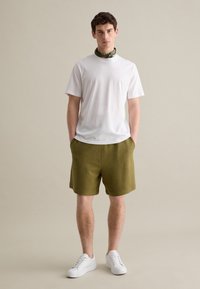 Weißes T-Shirt mit kurzen Ärmeln, olivgrüne Shorts und weiße Sneakers. Glattes Material, lockere Passform, mit einem Bandana um den Hals in grünem Muster.