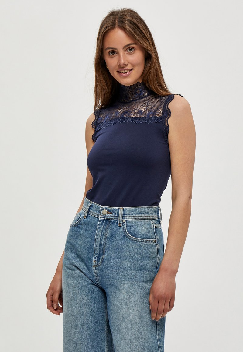 Marinefarbener Tanktop mit hohem Spitzenkragen und gefransten Rändern, kombiniert mit hoch-taillierten hellblauen Jeans. Glatte Stoffstruktur.