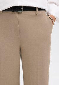 Pantaloni beige su misura con una texture liscia, caratterizzati da una parte frontale piatta, tasche laterali e una cintura nera con fibbia argentata.