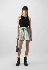 Débardeur noir ajusté, short rayé vert et blanc avec un nœud devant, casquette noire, collier en argent, bottes noires et sac en denim.
