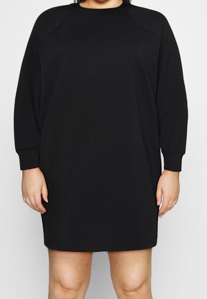 Robe sweat-shirt noire à manches longues et longueur au genou portée par une personne visible du cou à mi-cuisse sur un fond uni.