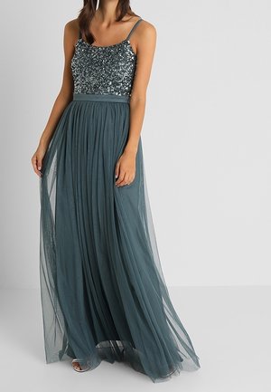 Vrouw die een mouwloze avondjurk draagt met een geborduurde lijfje en een vloeiende tule rok in gedempt teal, staand tegen een effen achtergrond.
