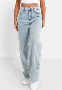 Donna che indossa jeans a gamba larga blu chiaro, top corto bianco e sneakers bianche, in piedi contro uno sfondo bianco uniforme.