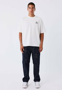 Camiseta blanca de manga corta con logo negro en el pecho, combinada con jeans de mezclilla azul oscuro con embellishments en forma de estrella y dobladillos doblados.