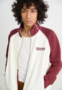 White and burgundy zip-up jacket with high collar, featuring branded embroidery. Worn over a white ribbed tank top. Smooth texture.