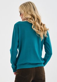 Kobieta z długimi kręconymi blond włosami, nosząca okulary przeciwsłoneczne, turkusowy sweter i brązowe kordowe spodnie, przedstawiona od tyłu na jednolitym tle.