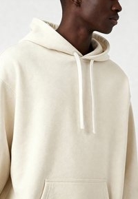 Asmuo, vilkintis kremiškai šviesią medvilninę džemperį su raišteliais ir priekine kišene, matomas nuo krūtinės iki viršaus ant vientiso fono.