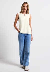 Blusa senza maniche color crema con dettaglio a chiave sul collo, abbinata a jeans a gamba larga di un leggero blu e sneakers bianche.