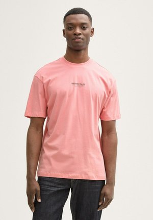 Camiseta de algodón rosa con cuello redondo, mangas cortas y un pequeño logo impreso en el pecho. Se llevan unos vaqueros de denim azul oscuro.