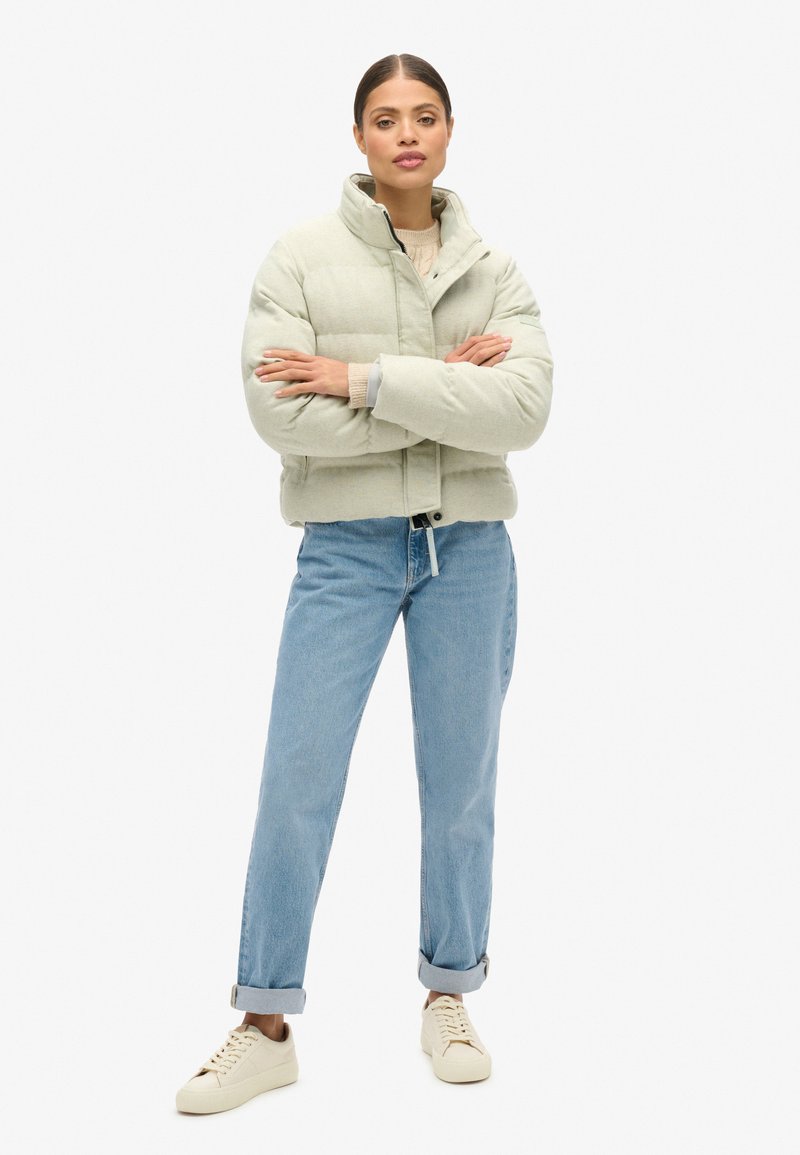 Dikke lichtgroene jas met een hoge kraag, voorzien van een sluiting aan de voorkant. Gecombineerd met opgerolde spijkerbroek en beige sneakers.