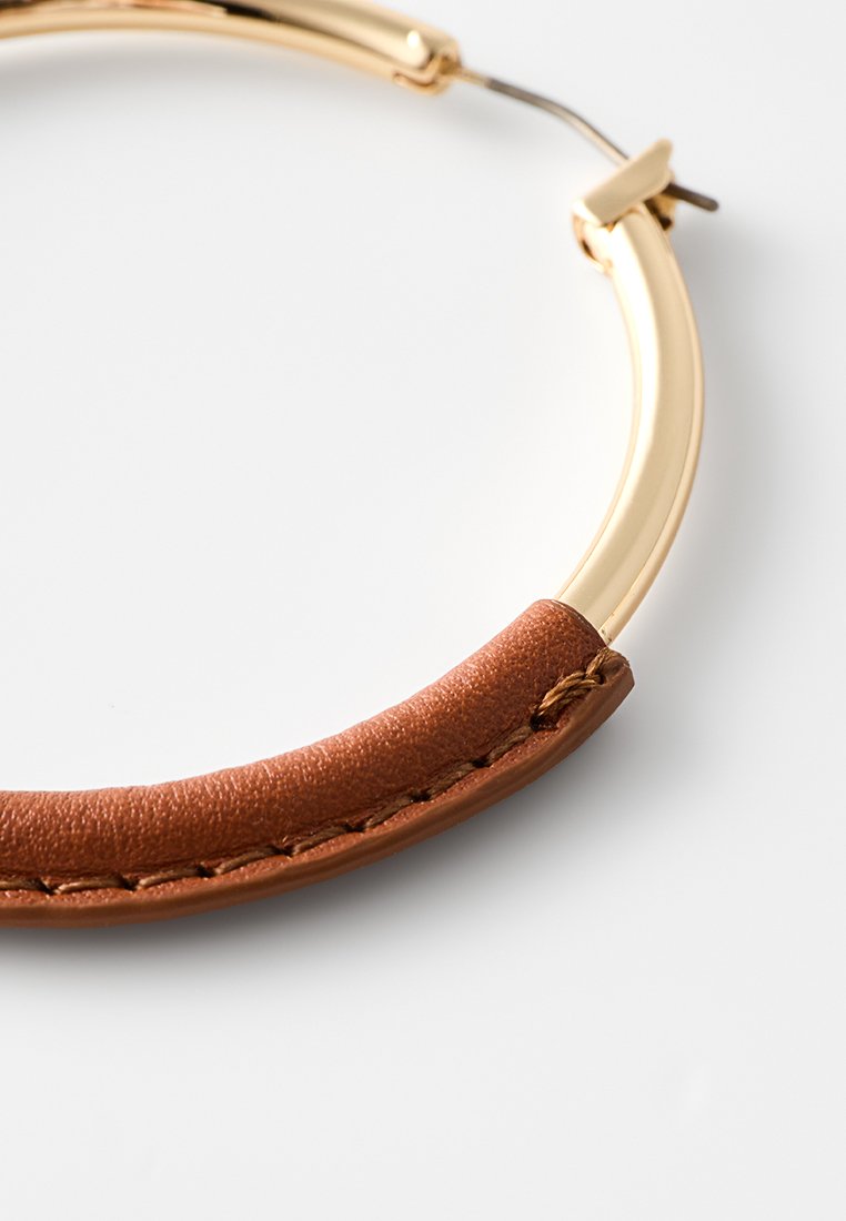 Boucle d'oreille en cuir marron avec un cadre en métal doré arrondi. Présente un accent en cuir cousu le long du bas de la boucle.