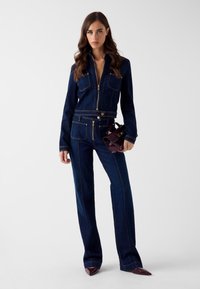 Kombinezon jeansowy z długimi rękawami, złotymi zamkami błyskawicznymi, kieszeniami na piersi i dopasowaną talią. Kolor ciemnoniebieski z kontrastowymi szwami.