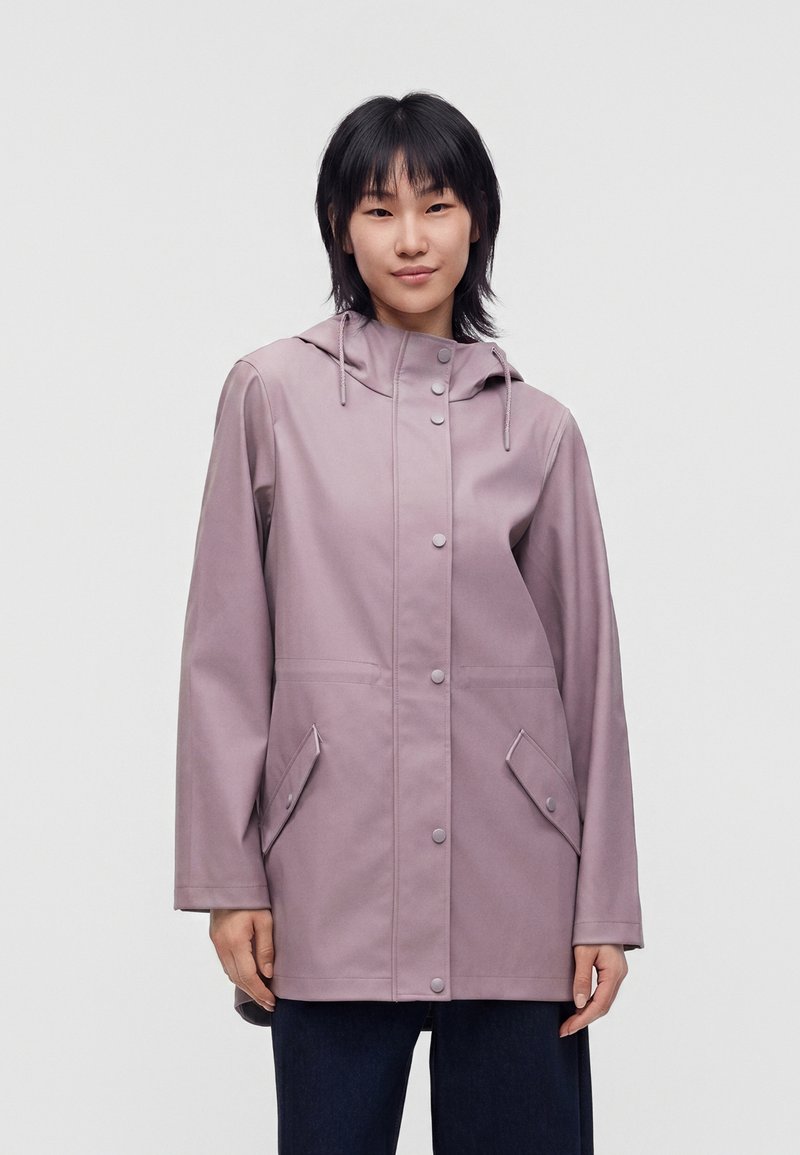 Jeune femme portant un imperméable à capuche violet clair avec des boutons-pression et des poches à rabat, debout devant un fond uni clair.