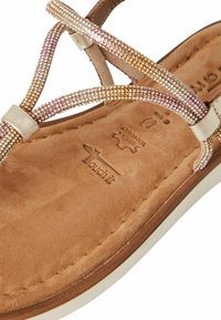 Sandale en daim marron avec des lanières entrecroisées et des accents métalliques, dotée d'une semelle contournée, ornée du nom et du logo de la marque.