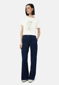 Wit cropped t-shirt met de tekst "JOIN THE PARTY" in zilver, gecombineerd met donkerblauwe flared jeans en lichtgroene schoenen.