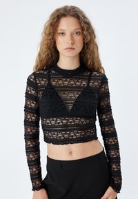 Sort blonderet top med lange ærmer og cropped design, der har intrikate mønstre og høj halsudskæring, parret med en ensfarvet sort nederdel.